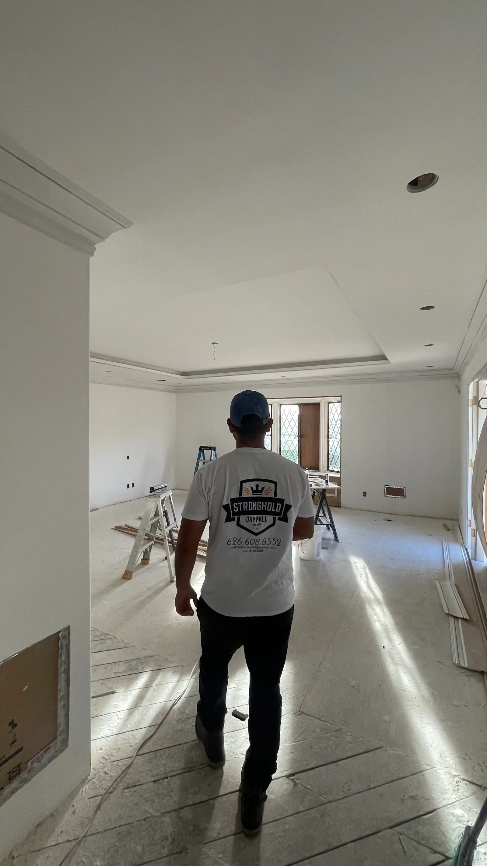 A Stronghold Drywall team member in a branded shirt walking through a finished bedroom, low sun raking across the floor and walls.