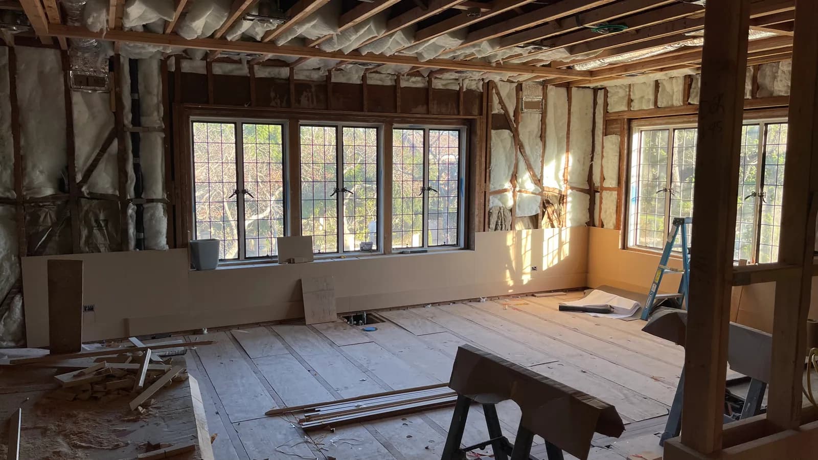 Open stud framing and ceiling joists in a large room with a row of original leaded-glass windows.