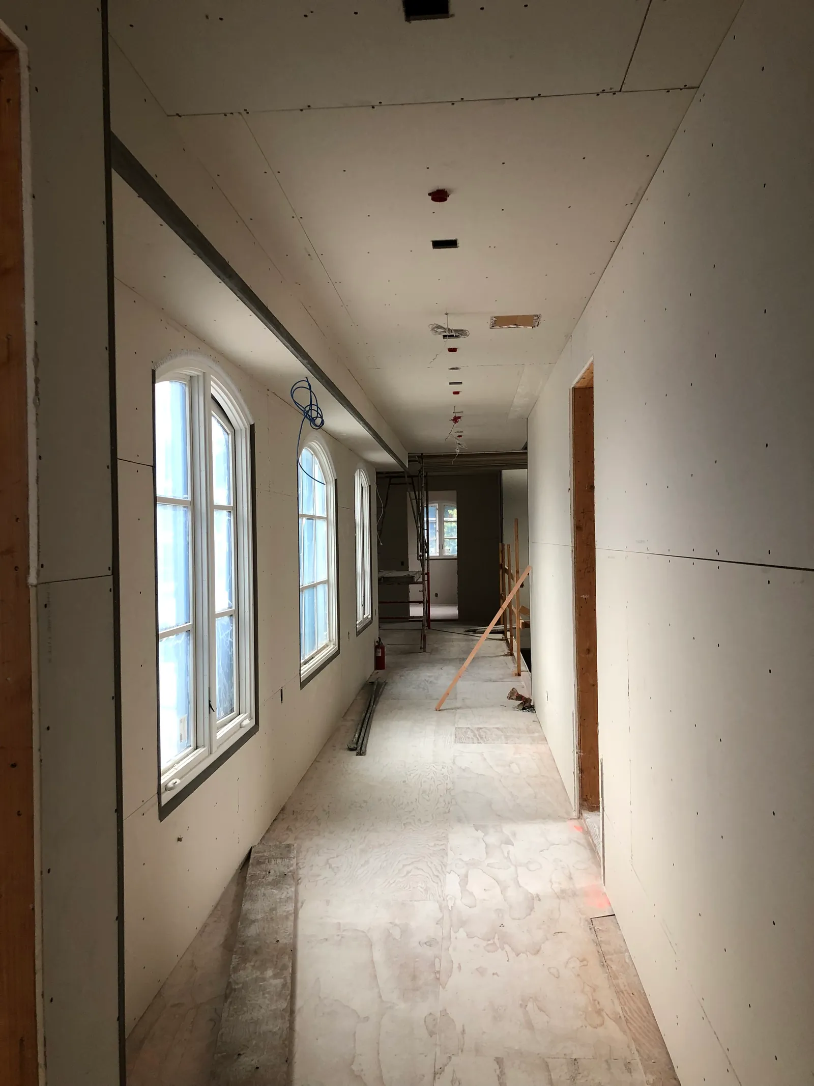 A long corridor with board hung end to end, seams and screw lines still visible, daylight entering from the far end.